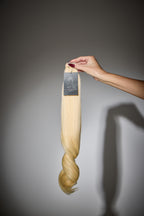 Blonde hair extension held by a hand against a gray background
